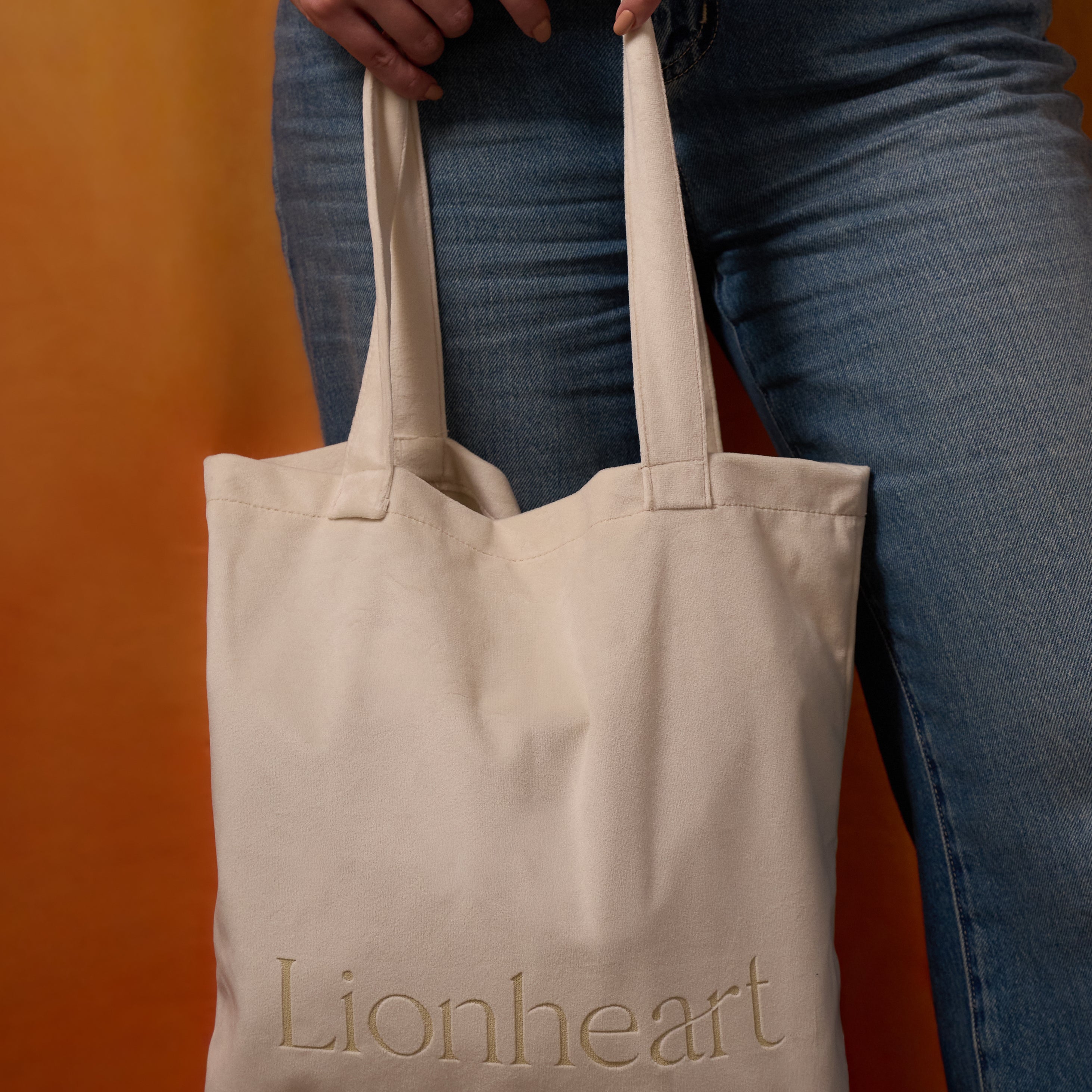 Beige tote bag with 'Lionheart' branding held by a person wearing blue jeans against an orange background