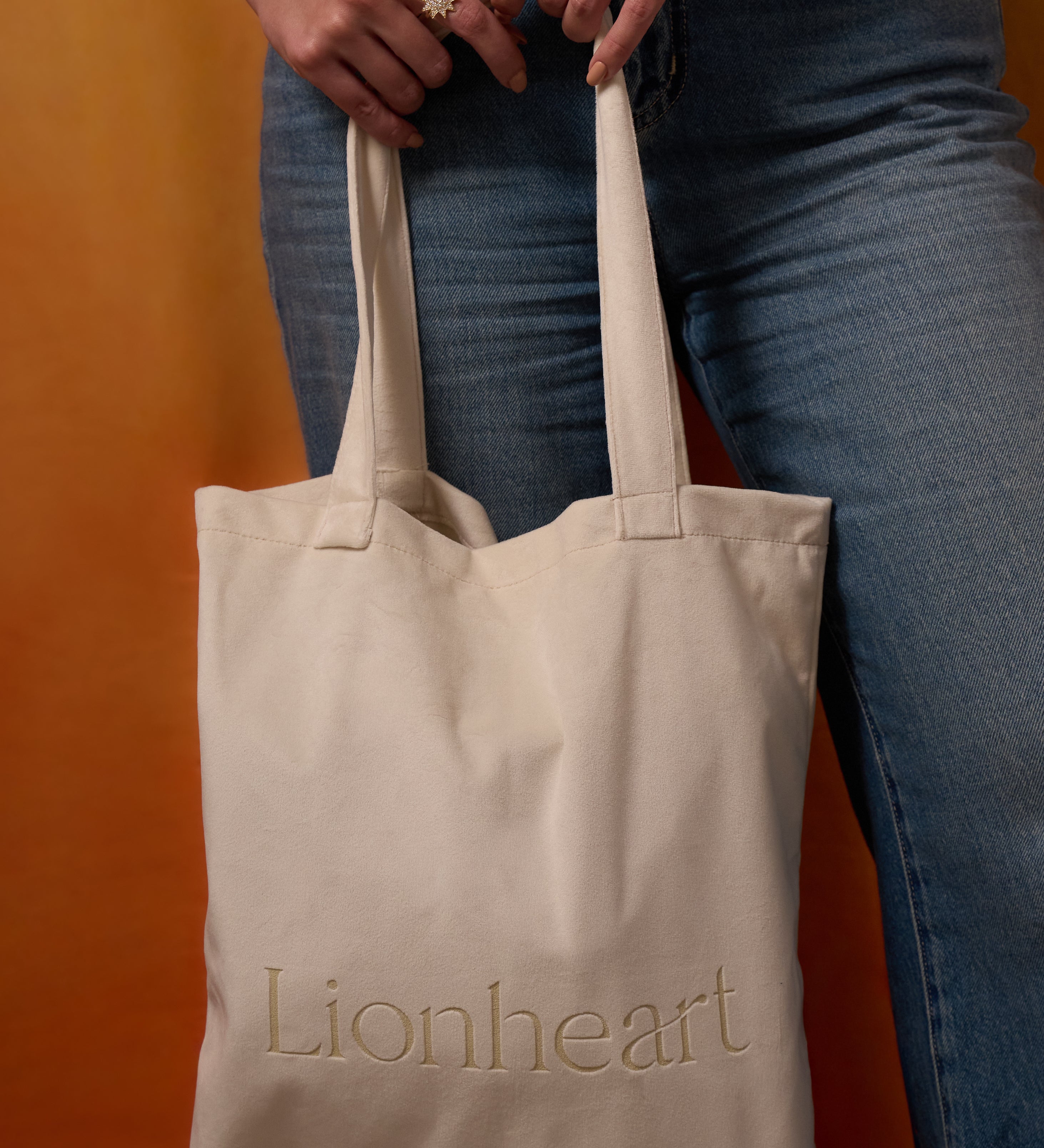 Beige tote bag with 'Lionheart' branding held by a person wearing blue jeans against an orange background