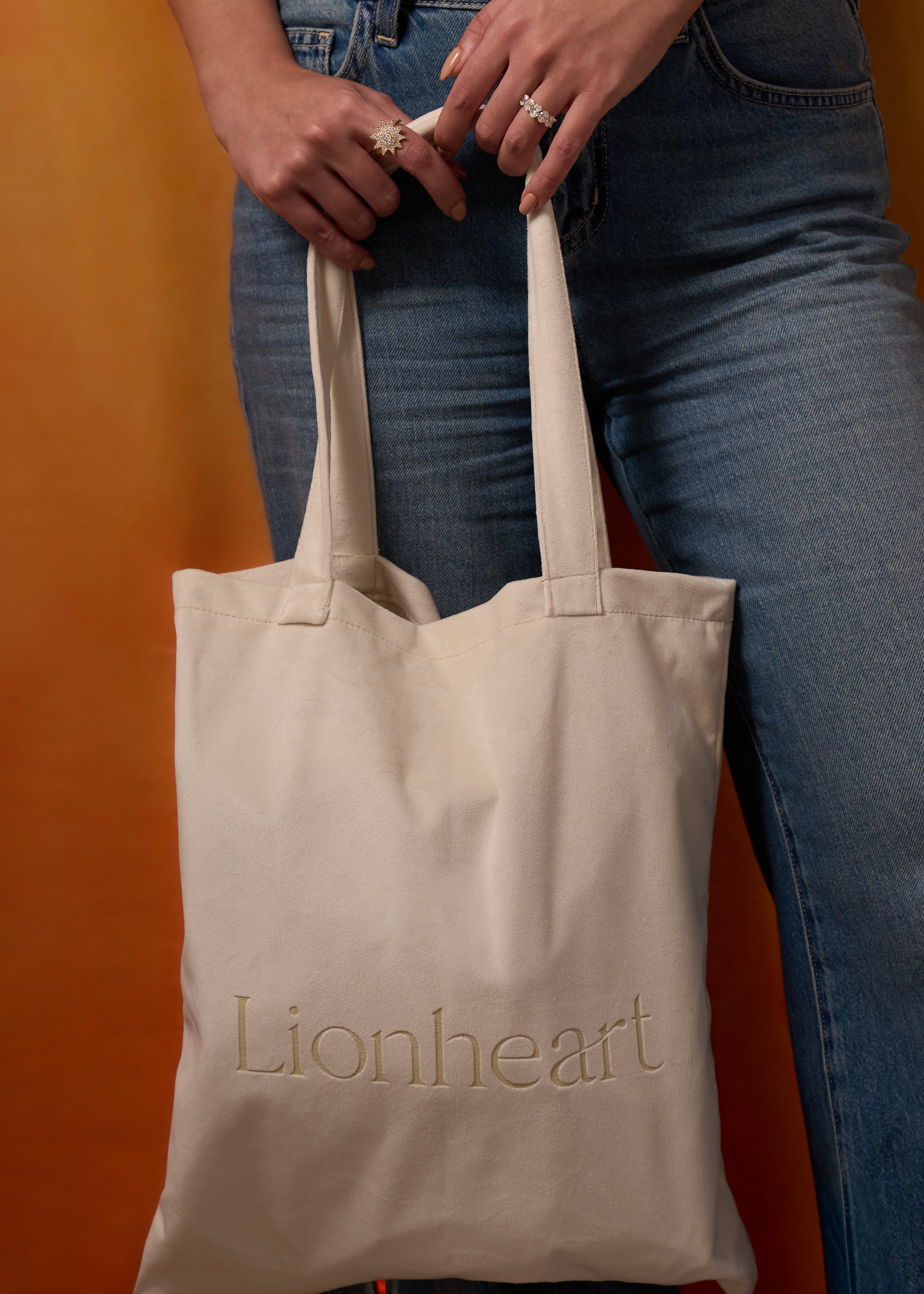 Beige tote bag with 'Lionheart' branding held by a person wearing blue jeans against an orange background