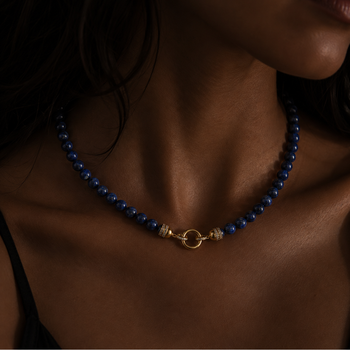 A close-up of a model wearing a beaded necklace with lapis beads and a gold clasp, set against a dark background.