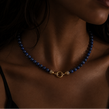 A close-up of a model wearing a beaded necklace with lapis beads and a gold clasp, set against a dark background.