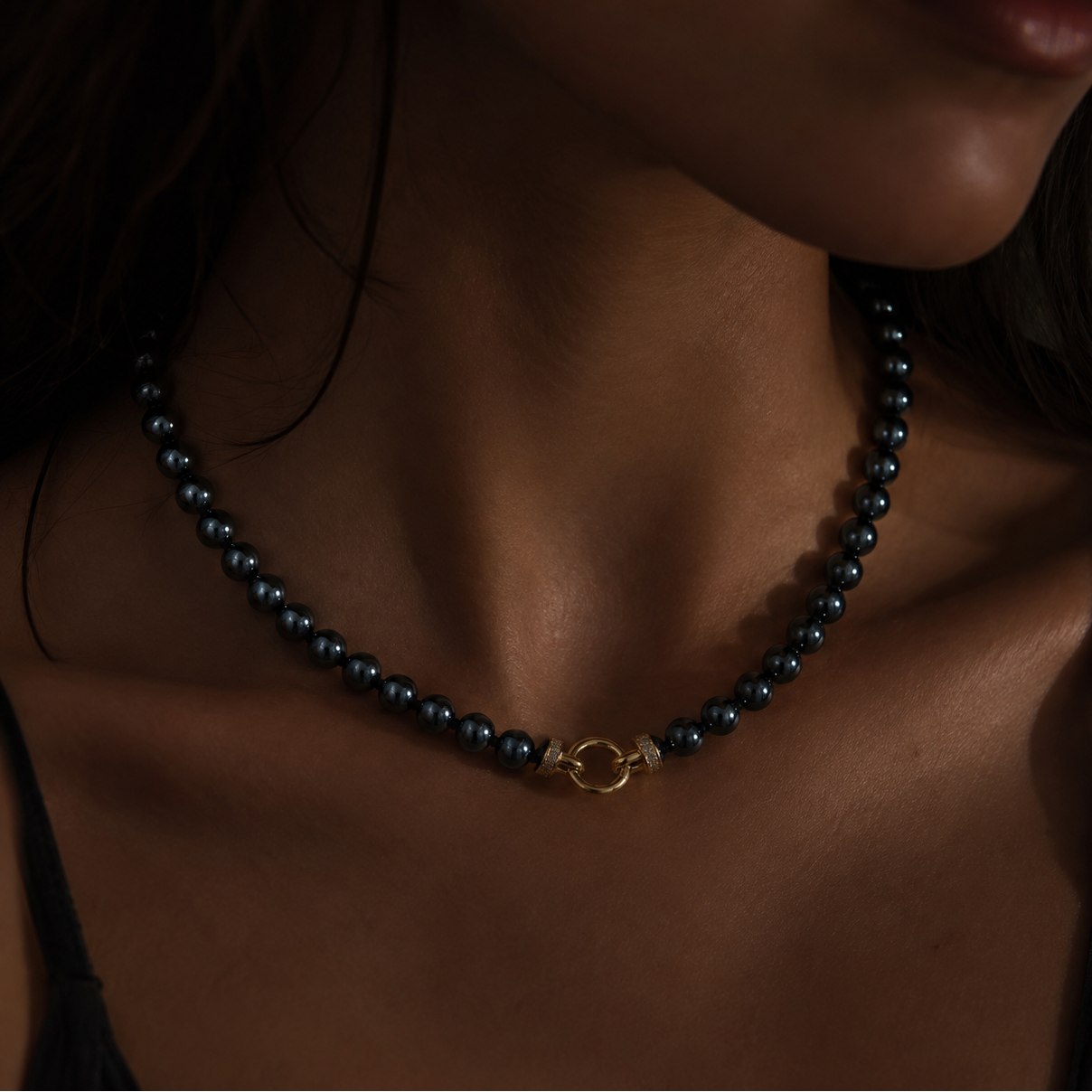 A close-up of a model's neck and chest wearing a hematite  beaded necklace with a small gold ring clasp.