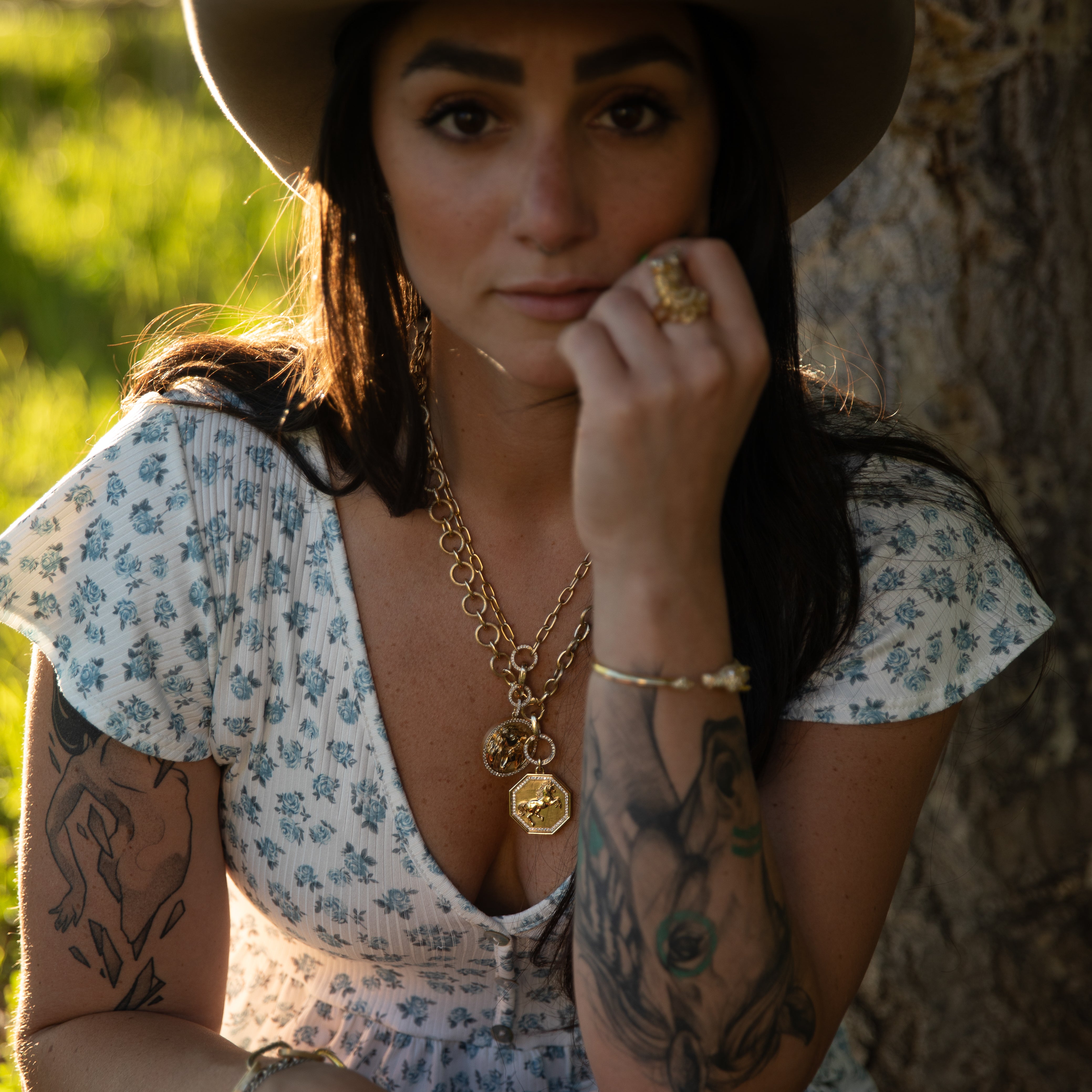 Woman in a floral outfit and cowboy hat with tattoos visible, sitting outdoors wearing Lionheart Jewelry 
